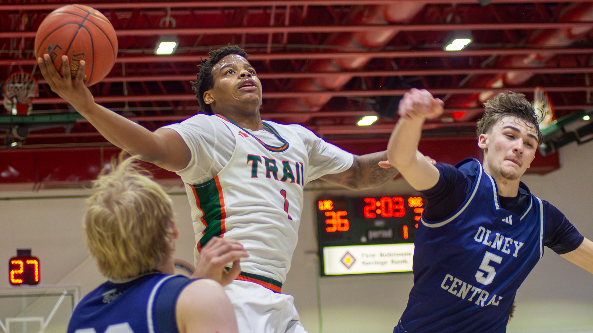 Mike White of Lincoln Trail College elevates toward the basket for a one-handed layup during a game, wearing a white Statesmen jersey with number 1. Two Olney Central defenders in blue contest the shot as the scoreboard and red ceiling beams are visible in the background.